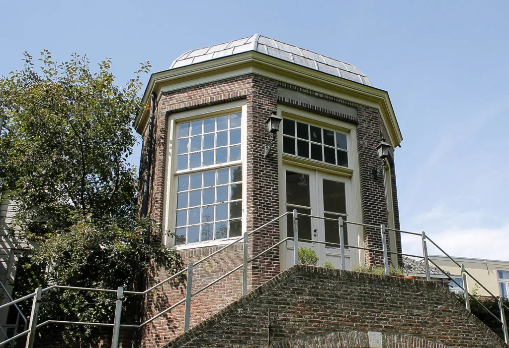 A small brick building with large windows and a dome-shaped roof, surrounded by greenery.