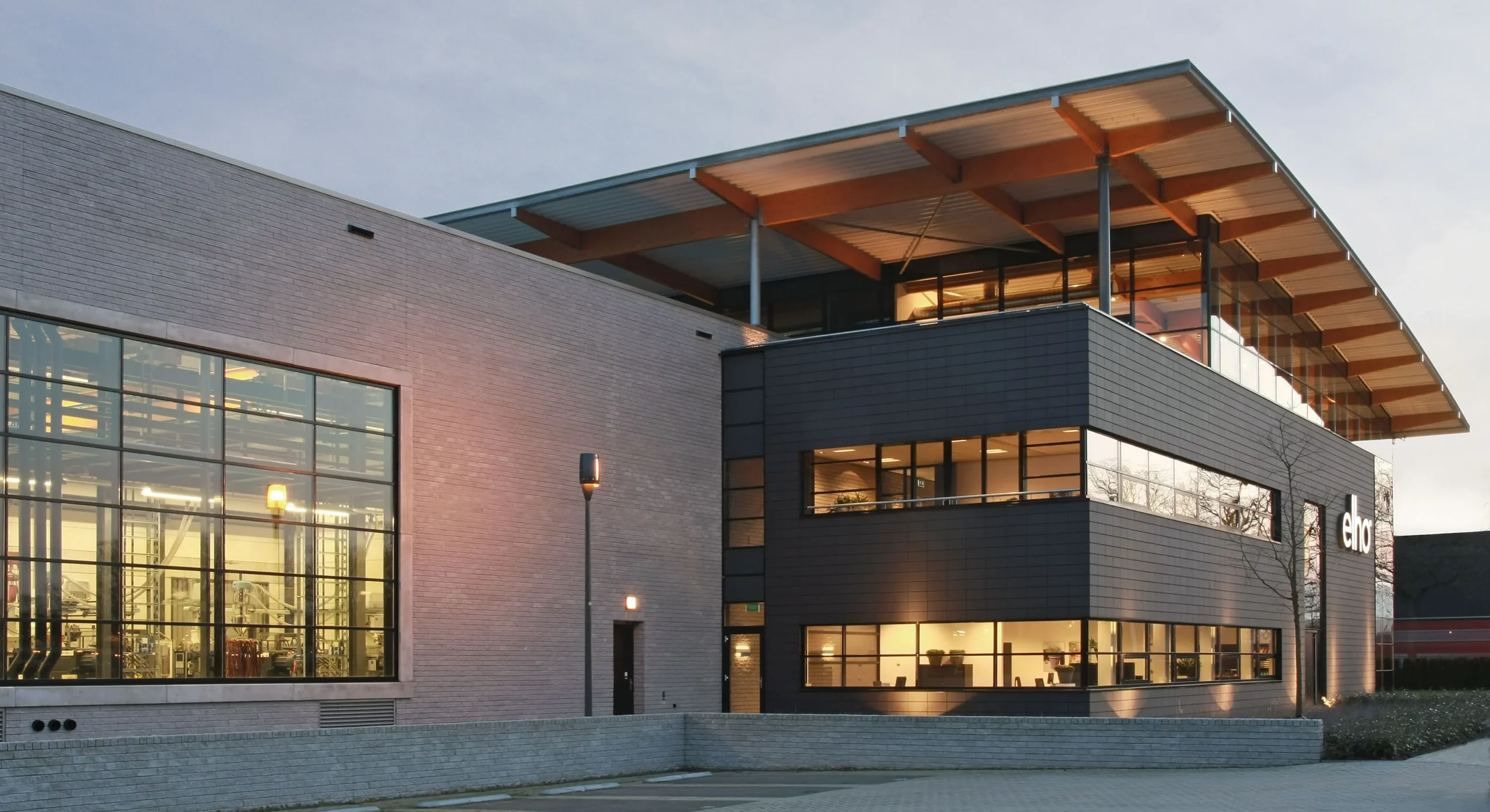 Modern building with large windows and a wooden roof structure at dusk, labeled "elho".
