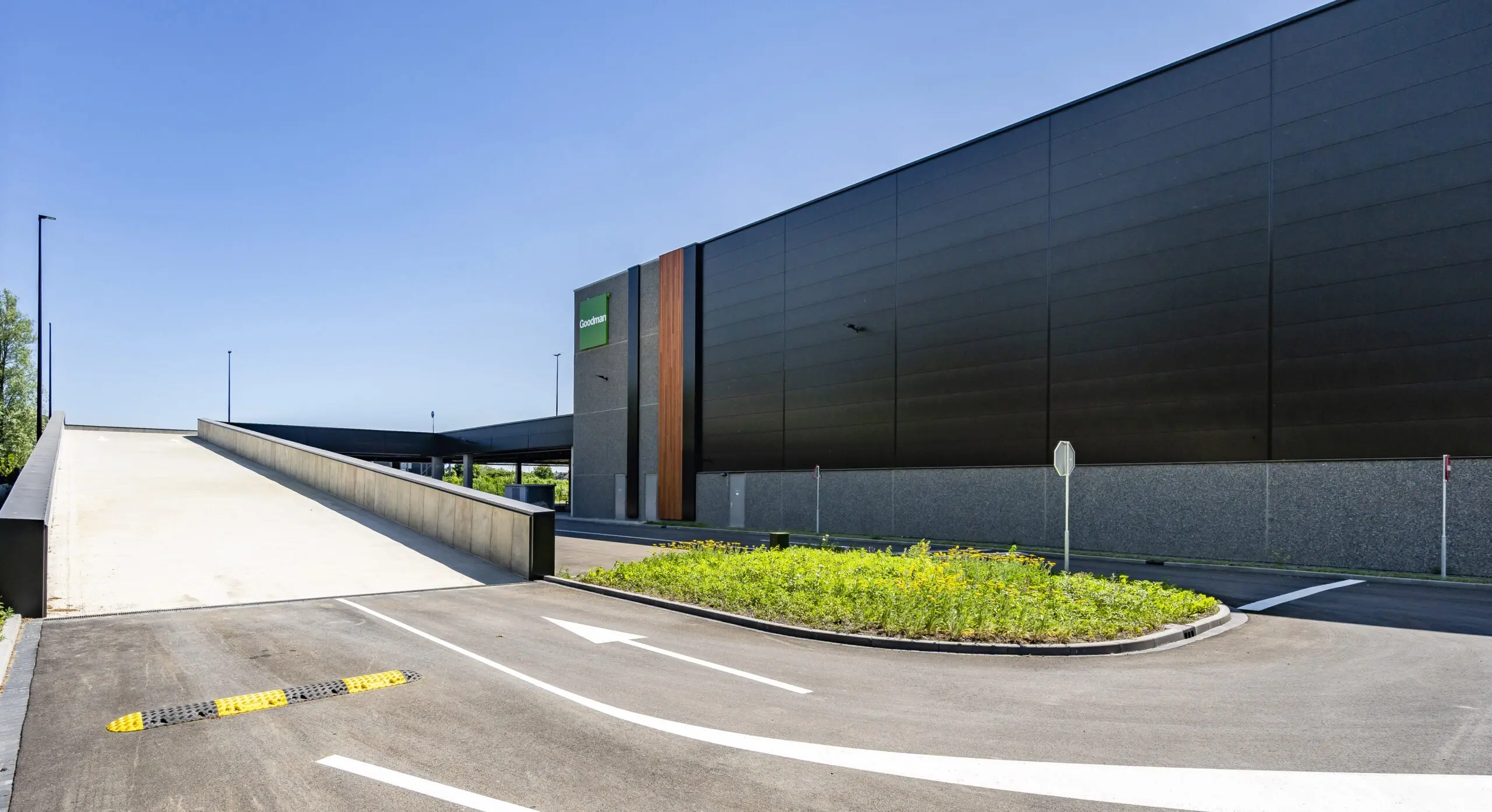 A modern warehouse building with a driveway and greenery under a clear blue sky.