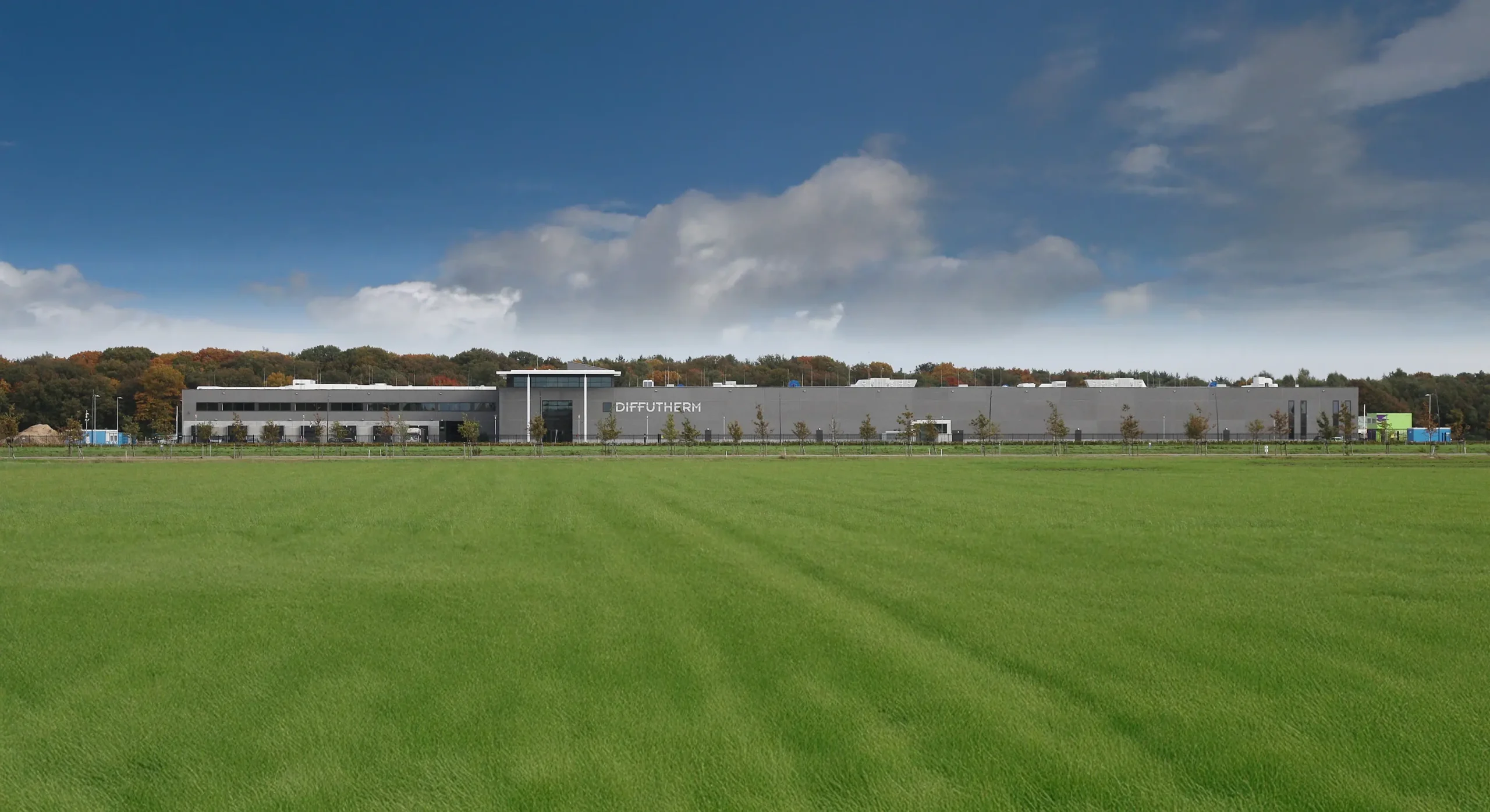 A modern industrial building labeled "Diffutherm" is situated behind a wide, green field under a partly cloudy sky.
