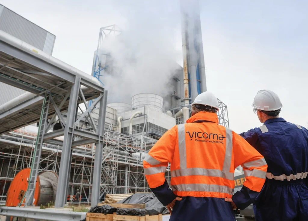 Two colleagues wearing safety gear look at an industrial installation emitting steam.