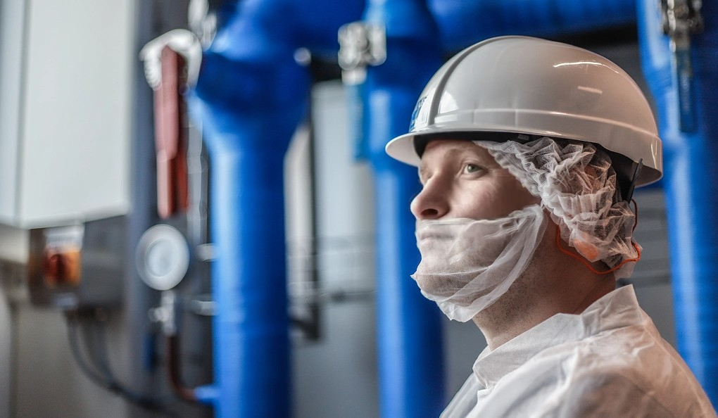 A man wearing a hard hat and face mask works in an industrial setting.