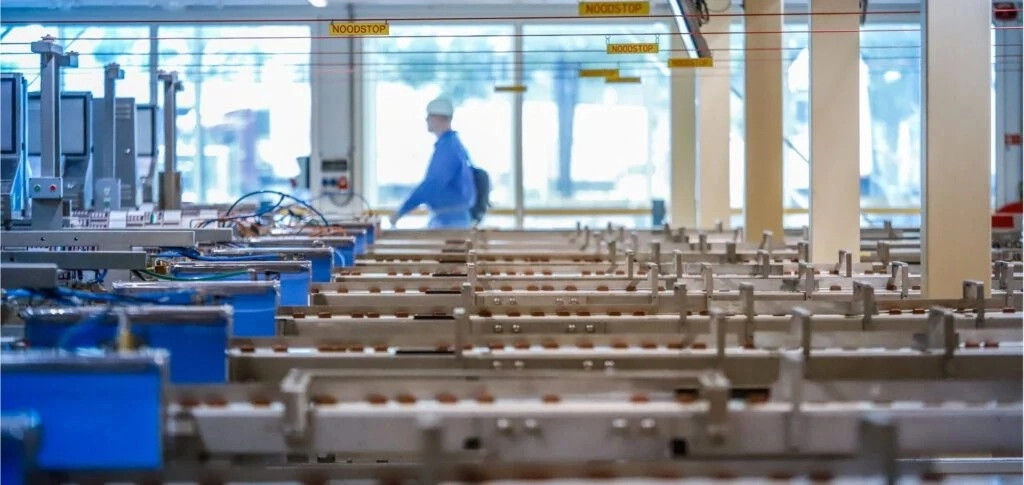 A factory worker in blue uniform walks past rows of automated machinery in an industrial setting.
