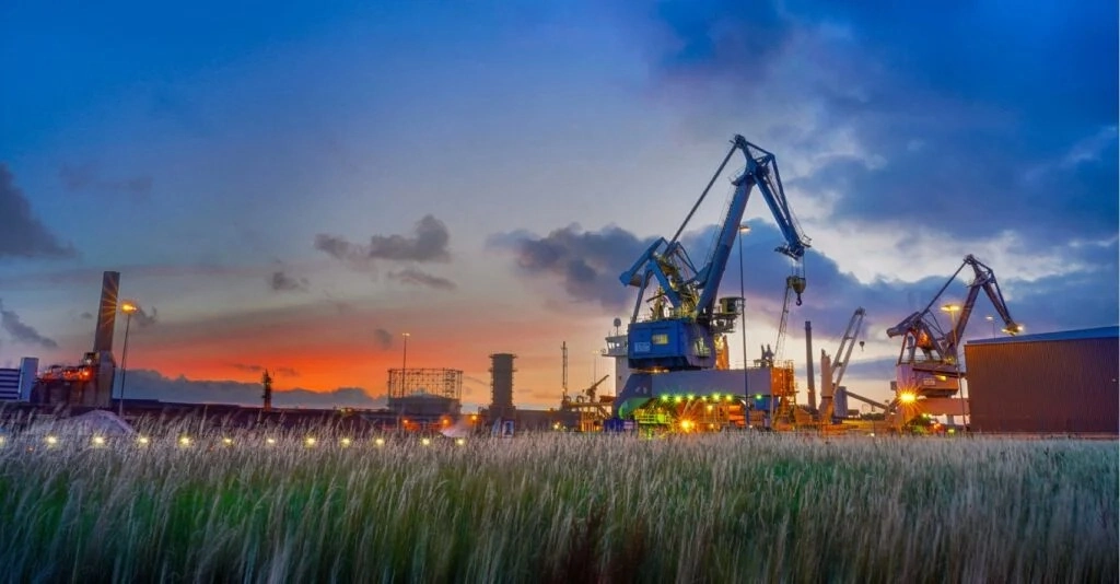 Industrial landscape with cranes and factory structures against a colorful sunset sky.