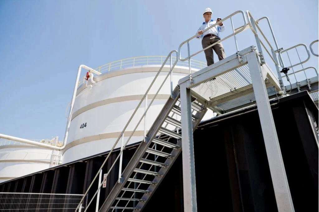 A person wearing a hard hat stands on a metal platform overlooking large industrial tank terminals.