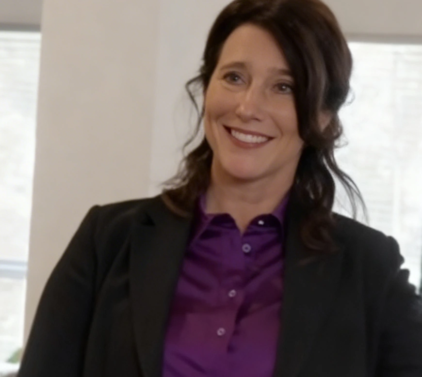 A woman in a purple blouse and black blazer smiling indoors.