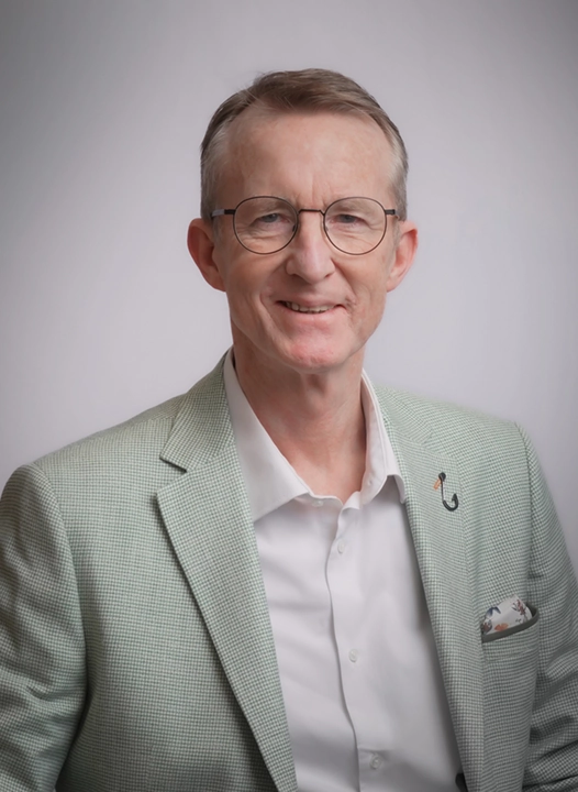 Een man met een bril en een licht geruit colbert poseert tegen een neutrale achtergrond.