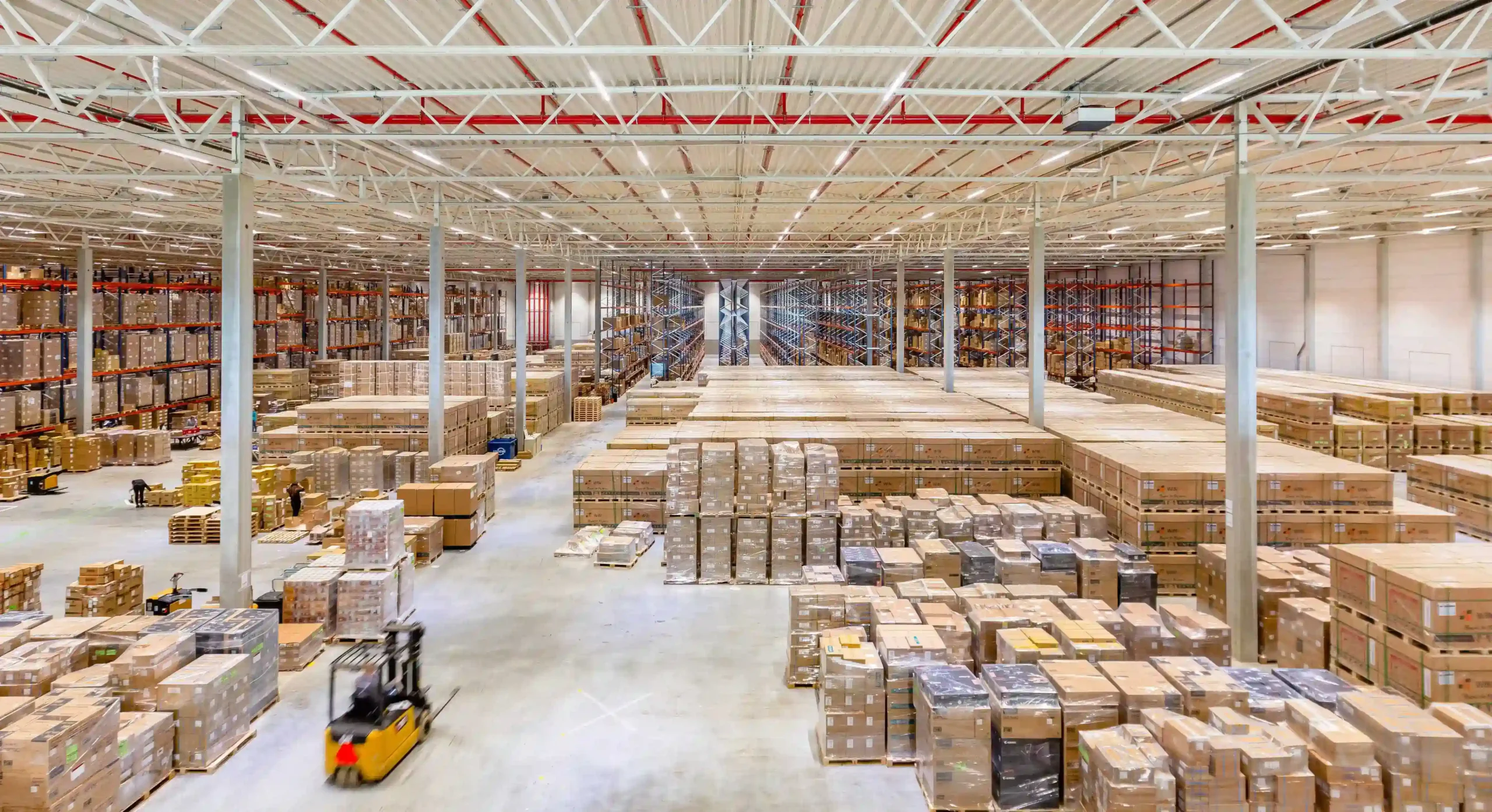 A spacious warehouse filled with stacked pallets and boxes, with a forklift moving in the foreground.