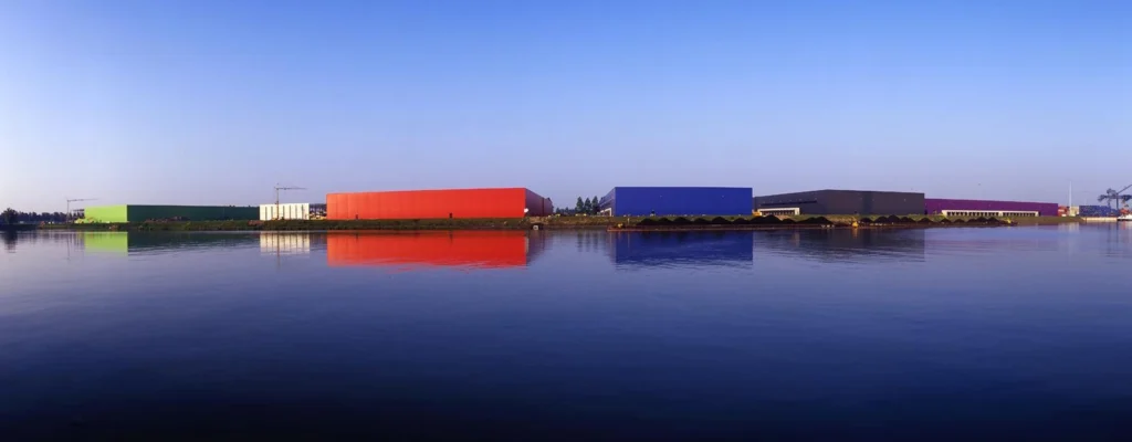 Colorful industrial buildings reflecting on the calm water of Seinehaven under a clear blue sky.