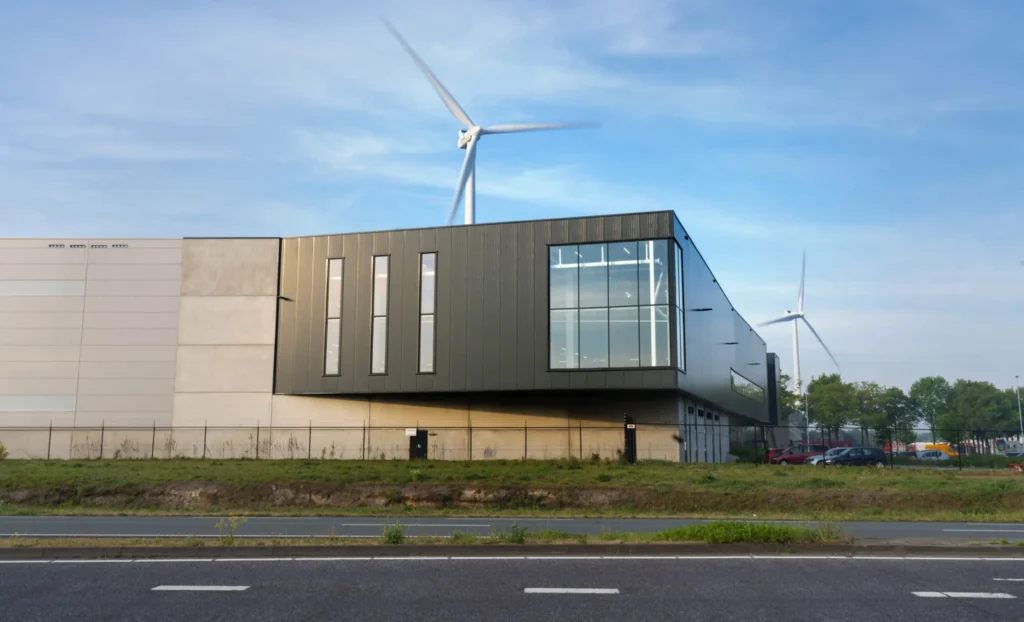 A modern industrial building with a wind turbine in the background under a blue sky.