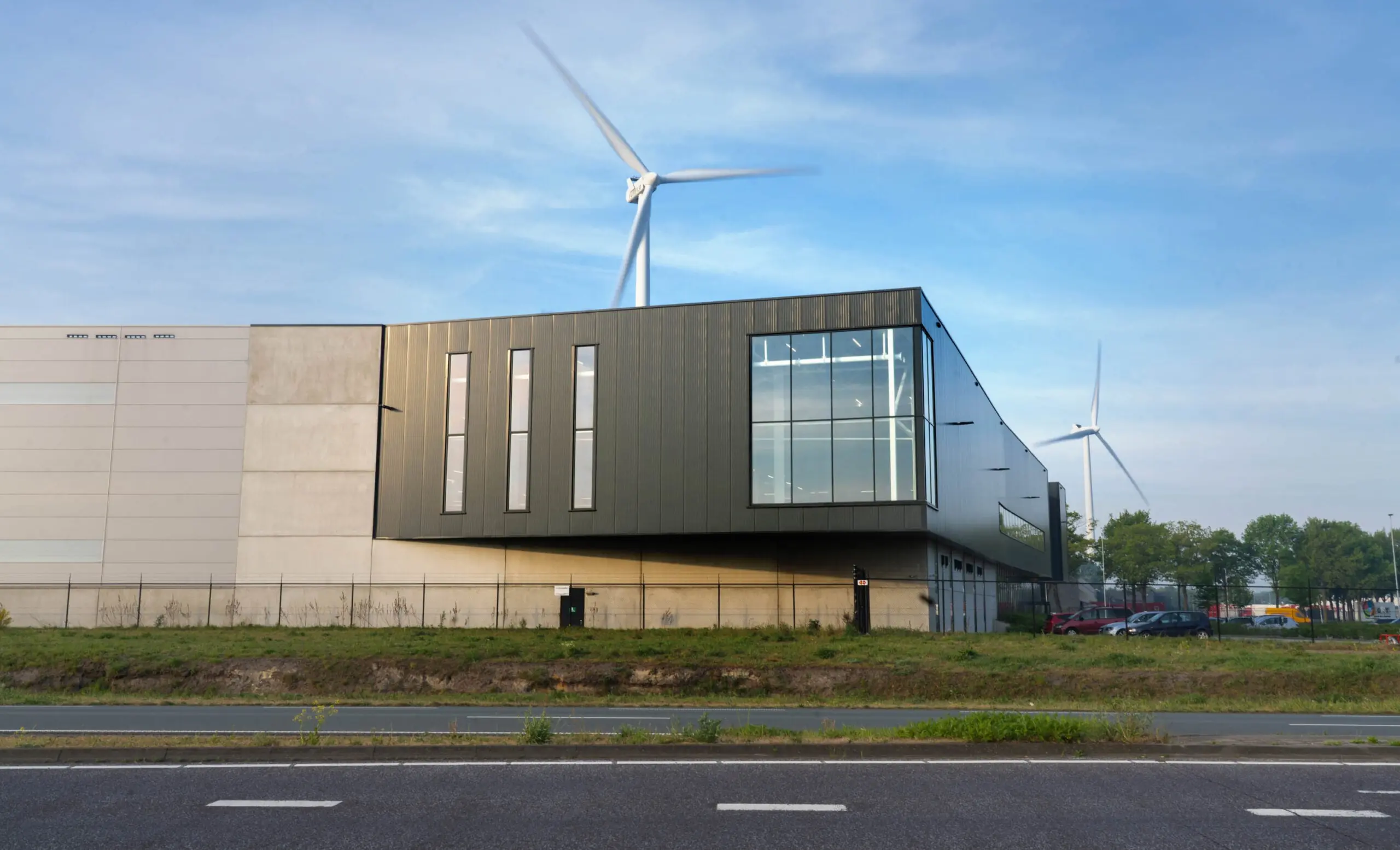 Een modern industrieel gebouw met grote ramen en een draaiende windturbine op de achtergrond.