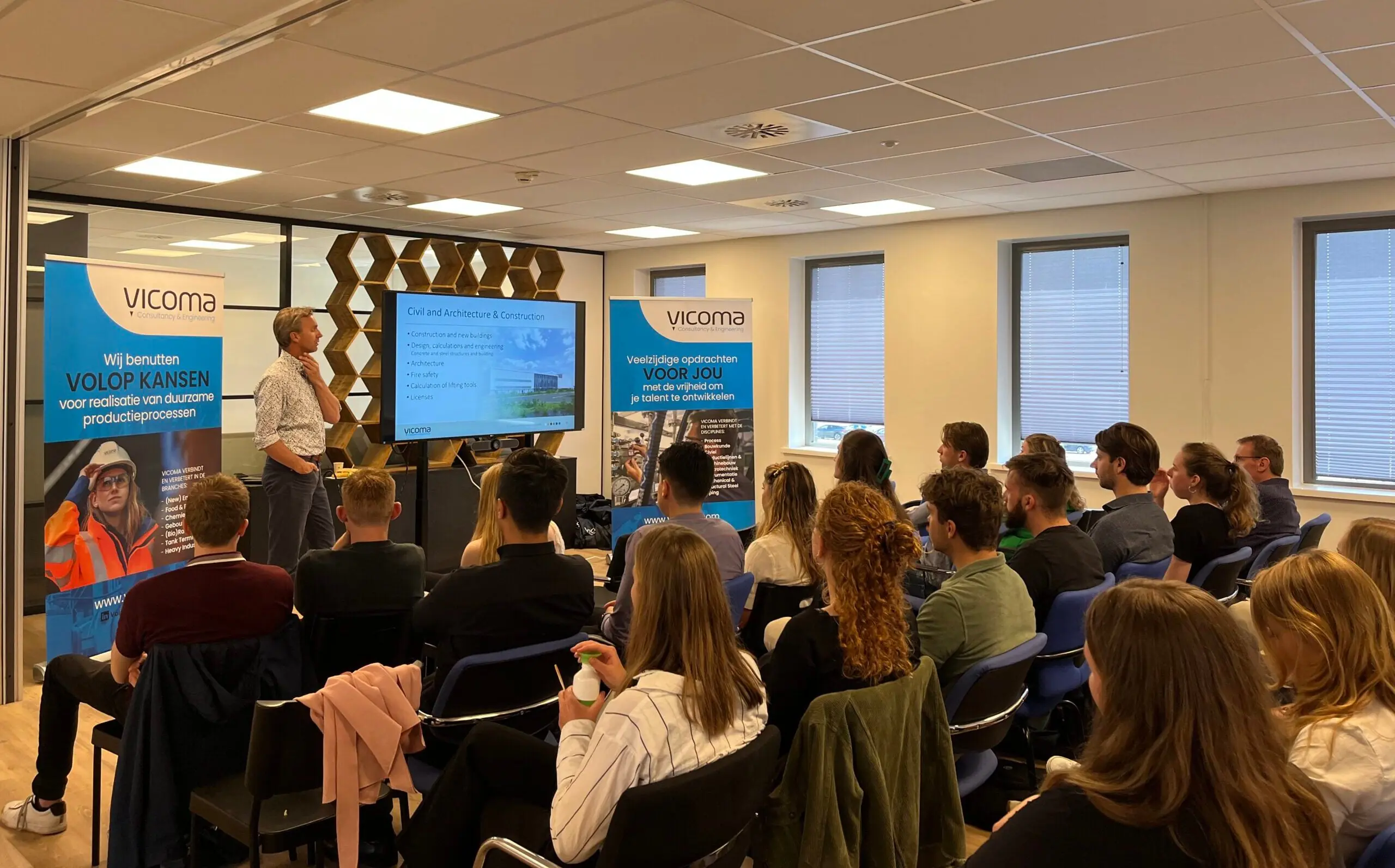 Een groep mensen in een lokaal luistert naar een presentatie van de Vicoma Groep over civiele en architectonische bouw.